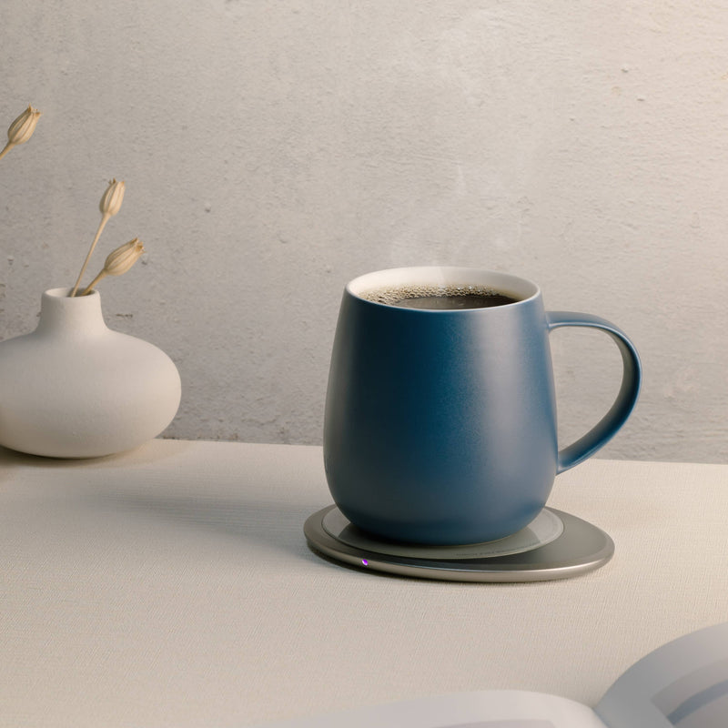 OHOM - Ui 3 Self-heating Mug Set: Jasmine White on warming coaster beside white vase, illustrating minimalist elegance and functionality with steaming coffee.