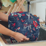 Hands rinse a navy-blue Bee's Wrap - Assorted 3 Pack - Botanical, featuring birds and floral motifs, under a kitchen faucet in a stainless-steel sink.