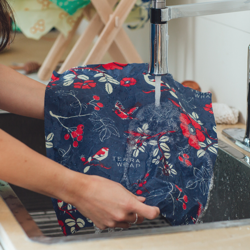 Hands rinse a navy-blue Bee's Wrap - Assorted 3 Pack - Botanical, featuring birds and floral motifs, under a kitchen faucet in a stainless-steel sink.