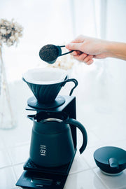 Hand scooping coffee grounds into a HARIO USA - V60 Metal Dripper, part of a precise pour-over setup with black server on digital scale.