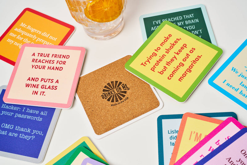 Drinks on Me coasters with funny one-liners scattered around a plain cork coaster. A citrus-garnished drink glass is visible above.