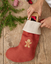 MagicLinen | Gingerbread Man Embroidered Christmas Stocking being filled with a gift box, accompanied by evergreen sprigs and festive twine on a wooden table.