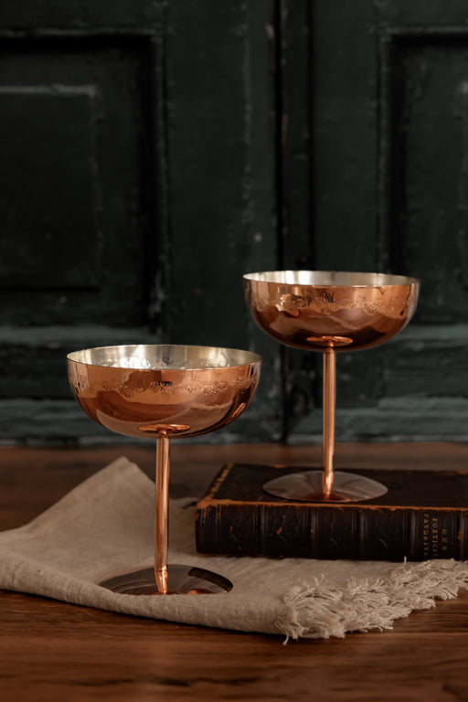 Coppermill Kitchen LLC | Vintage Inspired Copper Coupe Glass with floral engravings, displayed on linen and a book, exuding rustic elegance for upscale cocktail settings.