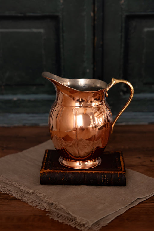 Coppermill Kitchen LLC | Vintage Inspired Large Copper Pitcher with brass-tone handle, atop a leather-bound book and linen cloth, showcasing elegant craftsmanship and vintage charm.