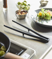 Yamazaki Home | Tower Floating Tweezer Tongs rest on a stainless-steel countertop among salad prep items like Brussels sprouts and salad, highlighting their drip-free floating design.