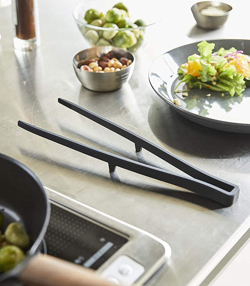 Yamazaki Home | Tower Floating Tweezer Tongs rest on a stainless-steel countertop among salad prep items like Brussels sprouts and salad, highlighting their drip-free floating design.