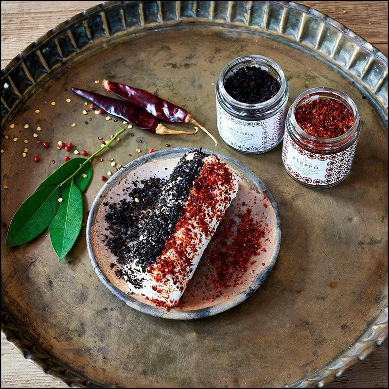 Villa Jerada - Urfa Biber - 50g spice jar on a tray with goat cheese rolled in Urfa Biber and Aleppo pepper, surrounded by garnishes.