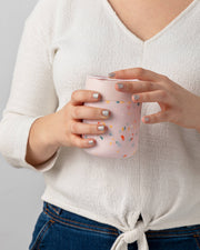 Hands with glittery nails holding a W&P - Reusable Ceramic Mug: Terrazzo Blush / 16 oz, featuring a matte silicone sleeve for a non-slip grip.