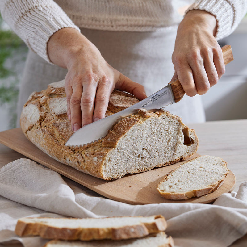 Hands slice a rustic loaf with the Opinel | Parallele N°116 Olivier Bread Knife, featuring a 21 cm stainless steel blade and olive wood handle.