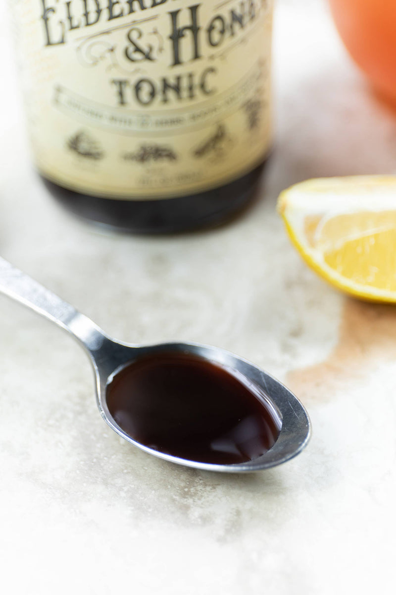 Roots And Leaves - Elderberry & Honey Tonic, 16 oz bottle, beside a spoon with liquid and lemon slice, highlighting herbal immune support features.