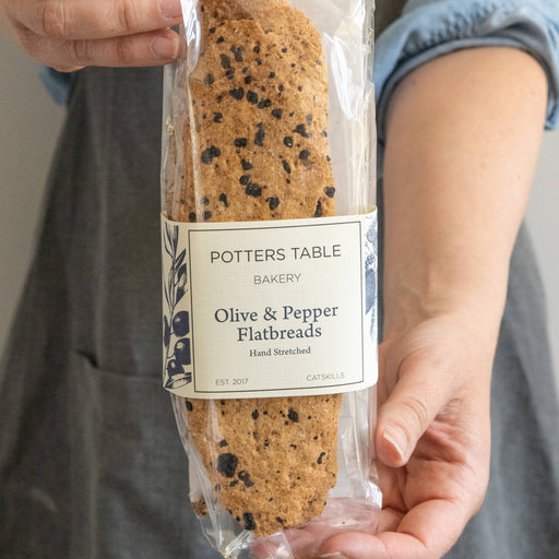 Potters Table - Cured Olive & Cracked Pepper Flatbreads, hand-stretched, visible olive specks, held in a clear bag, ideal for cheese spreads or charcuterie.