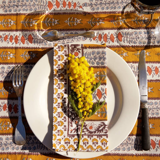 Avignon Napkins in Dijon & Blue adorn a rustic table setting with a white plate, mimosa sprig, and cutlery, embodying French elegance from Couleur Nature.
