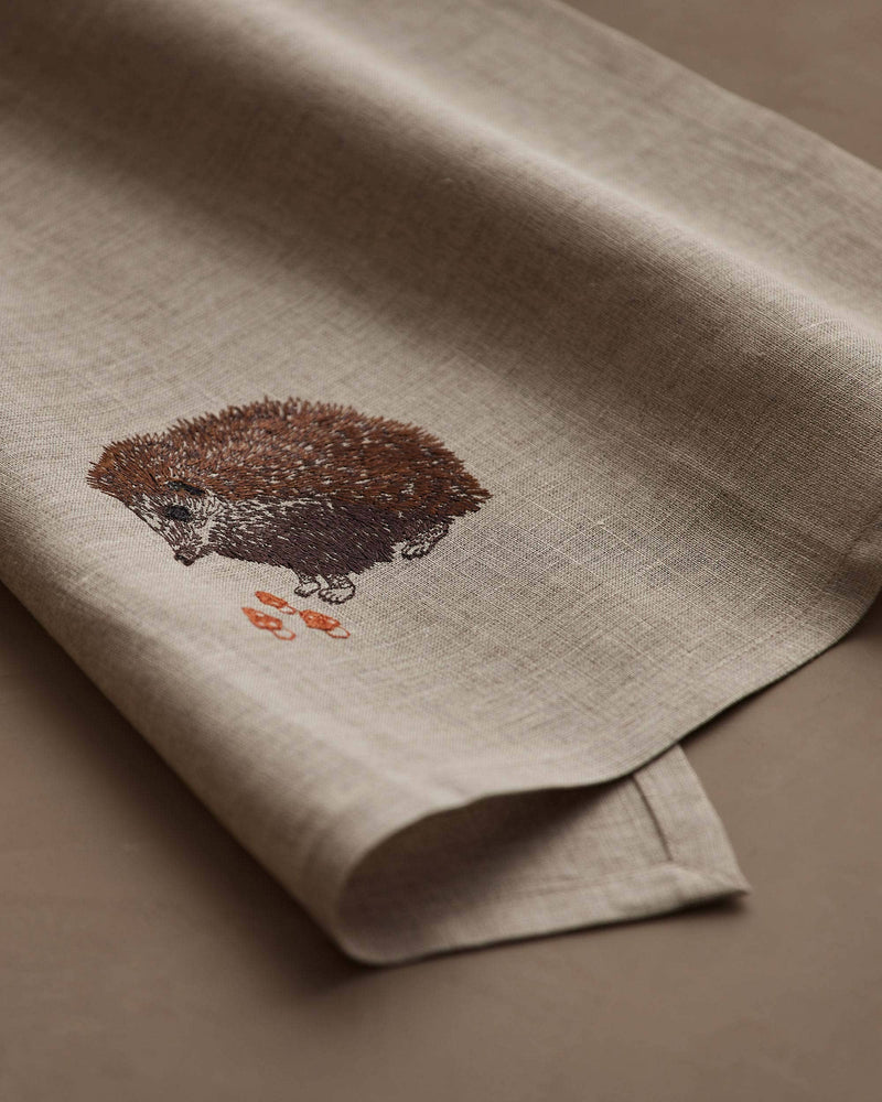 Coral & Tusk - Hedgehog with Mushrooms Tea Towel, featuring an embroidered hedgehog with orange mushrooms on natural linen, perfect for kitchen decor. Measures 25 × 18 unfolded.