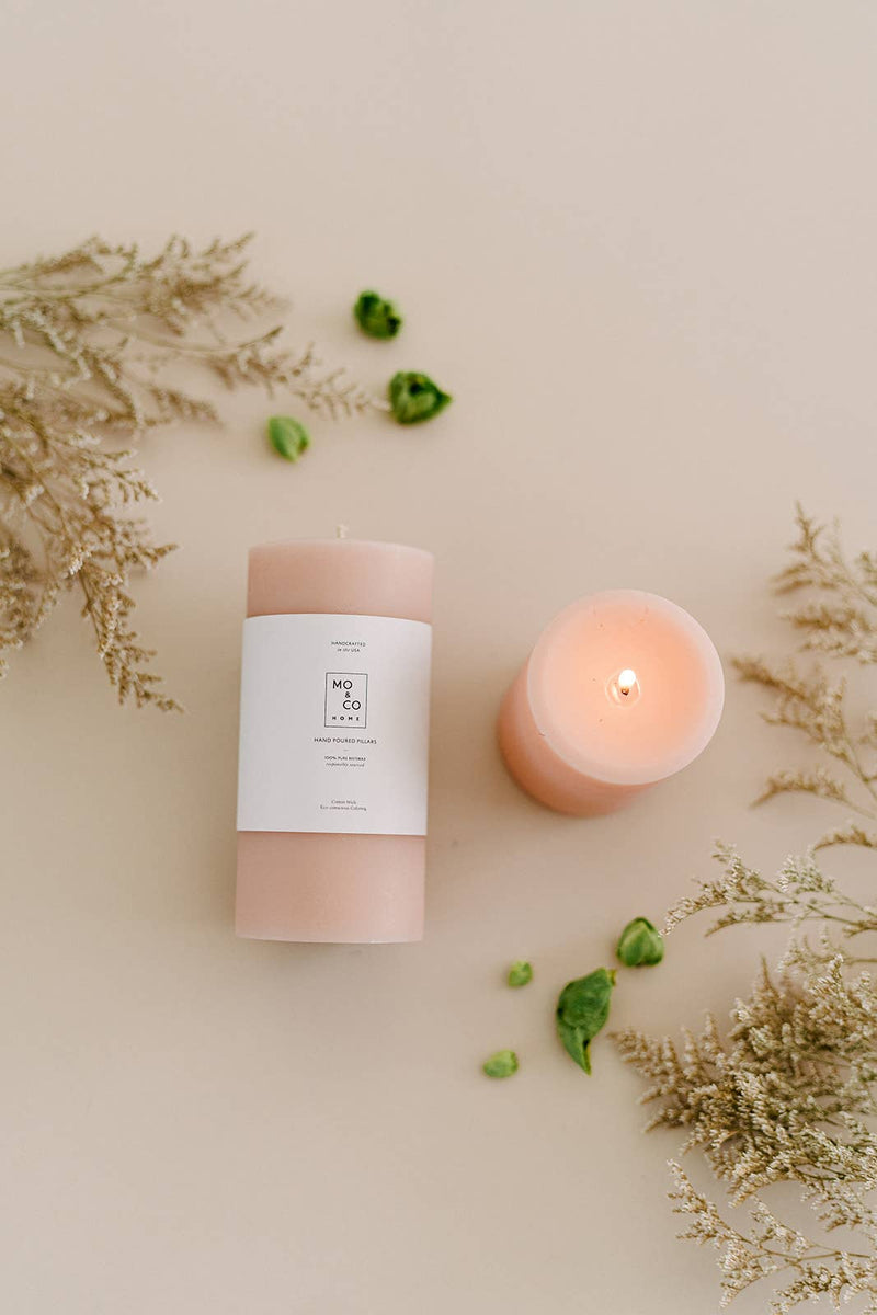 Mo&Co Home - 100% Beeswax Pillar Candles | Rose Pink: 3x6 Inch candle with a soft glow, displayed beside another candle, accompanied by green leaves. Perfect for home decor and celebrations.
