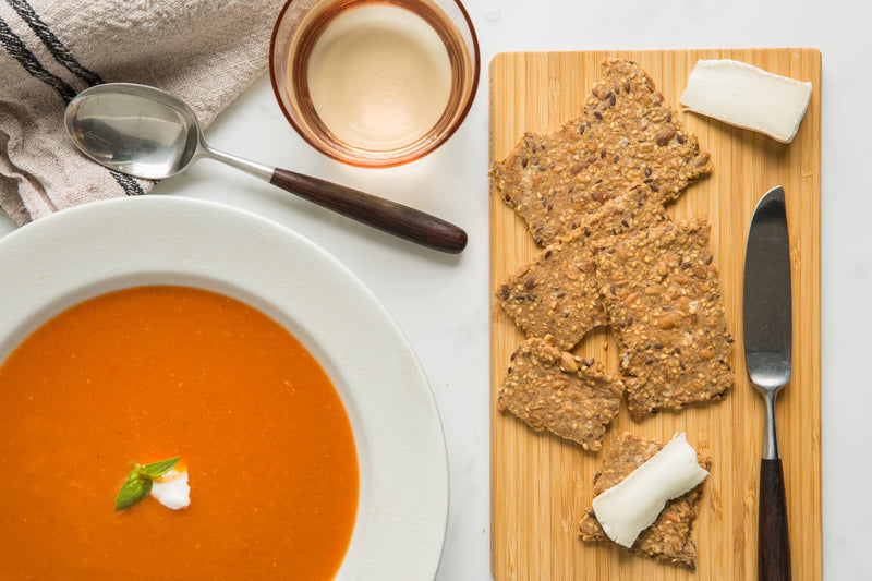 Norwegian Baked - Chili & Coriander Knekkebrød on a bamboo board, paired with cheese, complementing a tomato soup and drink in a curated lunch setting.