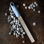 Microplane Premium Zester, Grater - Classic Blue, with stainless-steel blade and blue handle, shown on a rustic surface beside frozen blueberries. Perfect for zesting and grating tasks.