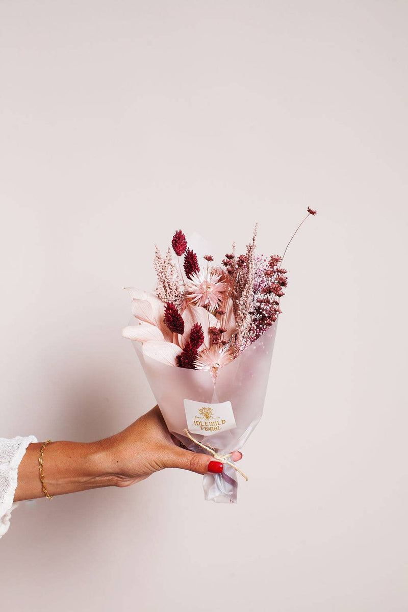 Idlewild Floral Co. - Sugarplum Dried Flower Bouquet, featuring preserved pink and burgundy blooms, held by a hand with red nails and a gold bracelet.