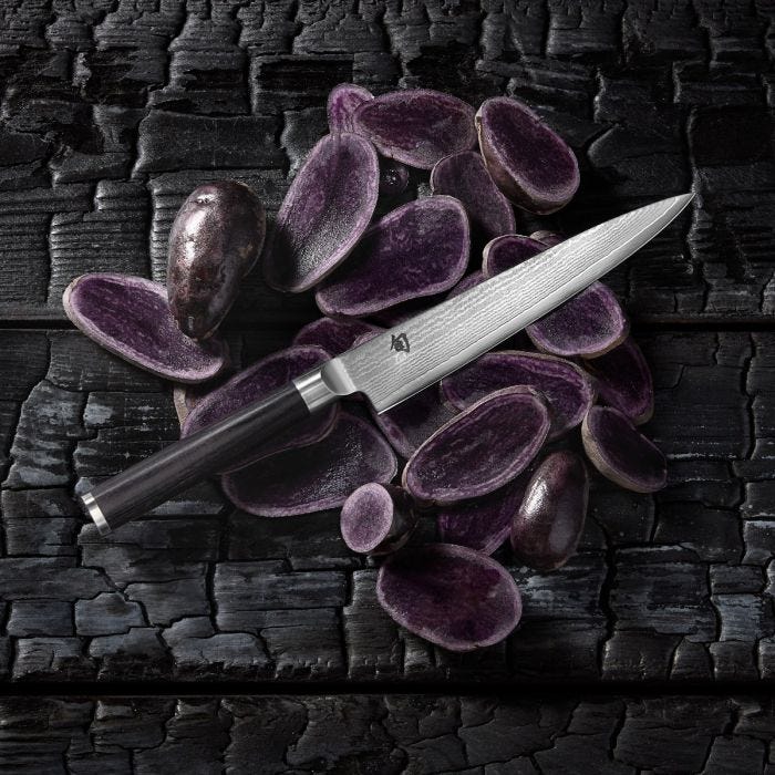 Shun Classic Utility Ebony, 6-inch knife with Damascus-pattern blade and dark wood handle on halved purple potatoes, showcasing precision and elegance on a charred wooden surface.