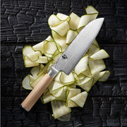 Shun Classic Blonde Santoku, 7, with a Damascus-pattern blade and light wooden handle, rests on thin vegetable ribbons atop a charred wood surface.