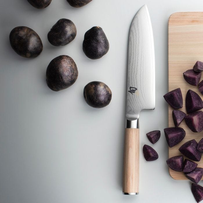 Shun Classic Blonde Santoku, 7 on a countertop, with wooden handle and Damascus-pattern blade, placed between whole and chopped purple potatoes on a cutting board.