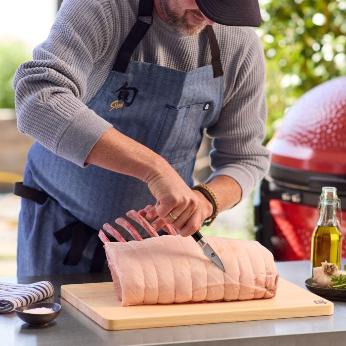 Shun Classic Bird's Beak Paring knife in use, trimming pork roast on a wooden board, surrounded by cooking essentials like salt, olive oil, and garlic.