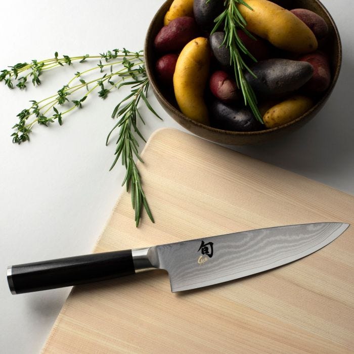 Shun | Classic Chef's Ebony, 6 inches knife on a wood cutting board with multi-colored fingerling potatoes and fresh herbs, showcasing its versatile culinary use.