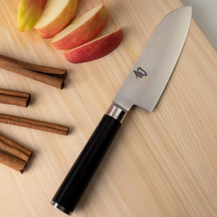 Shun | Classic Santoku Ebony, 5.5 inches, knife on a wooden board with apple slices and cinnamon sticks, highlighting its versatility for various kitchen tasks.