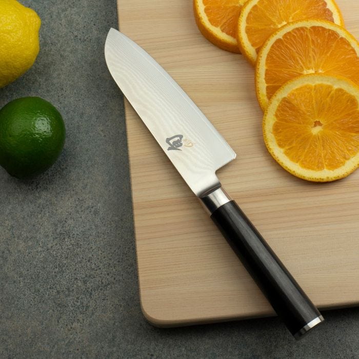 Shun | Classic Santoku Ebony, 5.5 inches rests on a wooden board with orange slices, showcasing its versatile design for medium kitchen tasks at Athens Cooks.