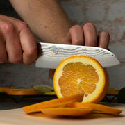 Hands slicing an orange with the Shun Classic Santoku Ebony, 5.5 inches knife on a cutting board, showcasing its precision and craftsmanship.