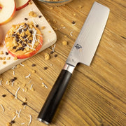 Shun Classic Nakiri Ebony, 6.5 inches, rests on a wooden countertop, surrounded by apple slices, nut spread, granola, coconut, and chocolate chips, showcasing its vegetable preparation prowess.