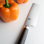 Shun Classic Nakiri Ebony, 6.5 inches, next to two orange bell peppers on a smooth surface, showcasing its straight blade and elegant design for vegetable preparation.