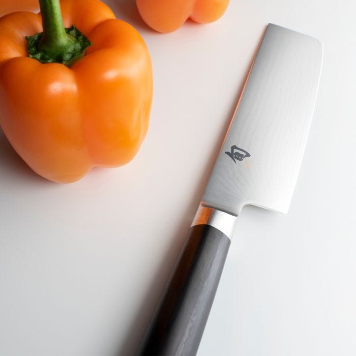 Shun Classic Nakiri Ebony, 6.5 inches, next to two orange bell peppers on a smooth surface, showcasing its straight blade and elegant design for vegetable preparation.