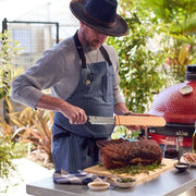 Man slicing roast with Shun Classic Hollow Ground Brisket Knife beside red kamado grill, highlighting precision cutlery from Athens Cooks.