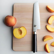 Shun | Classic Master Utility Ebony, 6.5, displayed on a cutting board with peaches, highlighting its versatile chef's knife features ideal for precise food preparation.