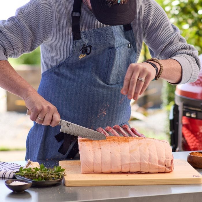 Person in denim apron uses Shun Classic Blonde Master Utility, 6.5 knife to season and slice rib roast on wooden board, with herbs and spices nearby.