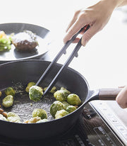 Yamazaki Home | Tower Floating Tweezer Tongs, Silicone, used to pick up Brussels sprouts from a skillet on an induction cooktop, showcasing their drip-saving floating design.
