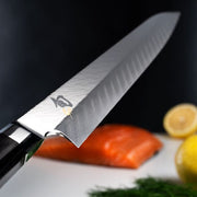 Shun Dual Core Yanagiba knife glides above a cutting board with salmon, lemons, and herbs, showcasing its precision blade and sushi-cutting capability.