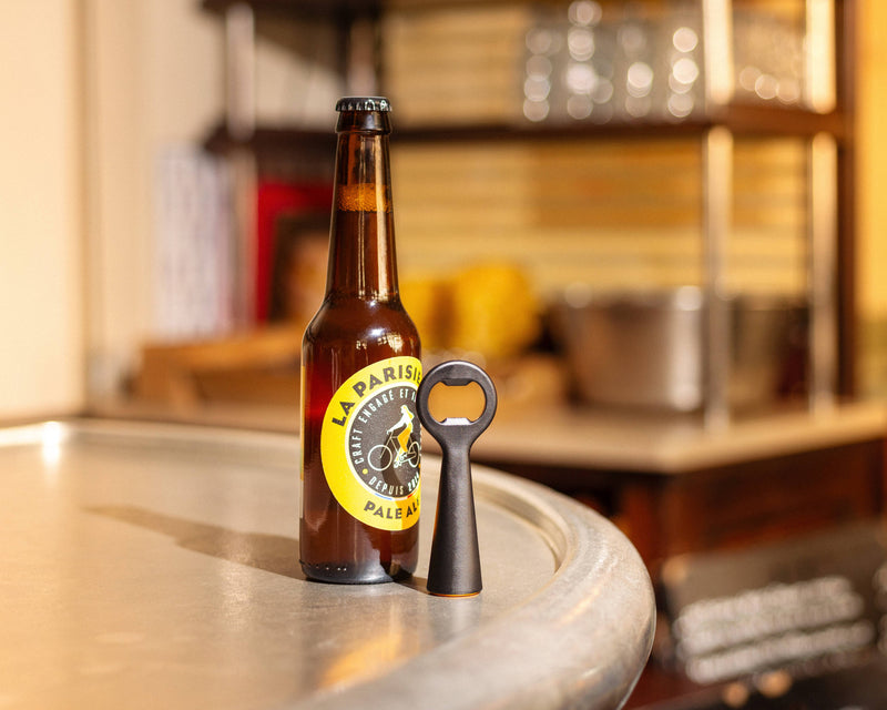 L'Atelier du Vin - USA - Bottle Opener - LE BRASSEUR – BLACK stands upright beside a La Parisienne Pale Ale bottle on a bar-top.