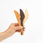 Hand holding three Collin Garrity wooden spreaders in light, medium, and dark wood, ideal for cheese, jam, and butter, against a white background.