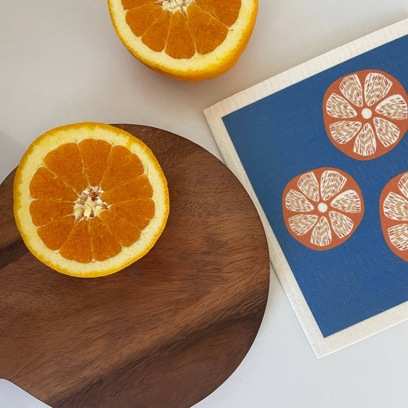 Ink and Fiber Designs - Oranges Swedish Dishcloth on a wooden board, featuring vibrant orange patterns, ideal for eco-friendly kitchen cleaning.