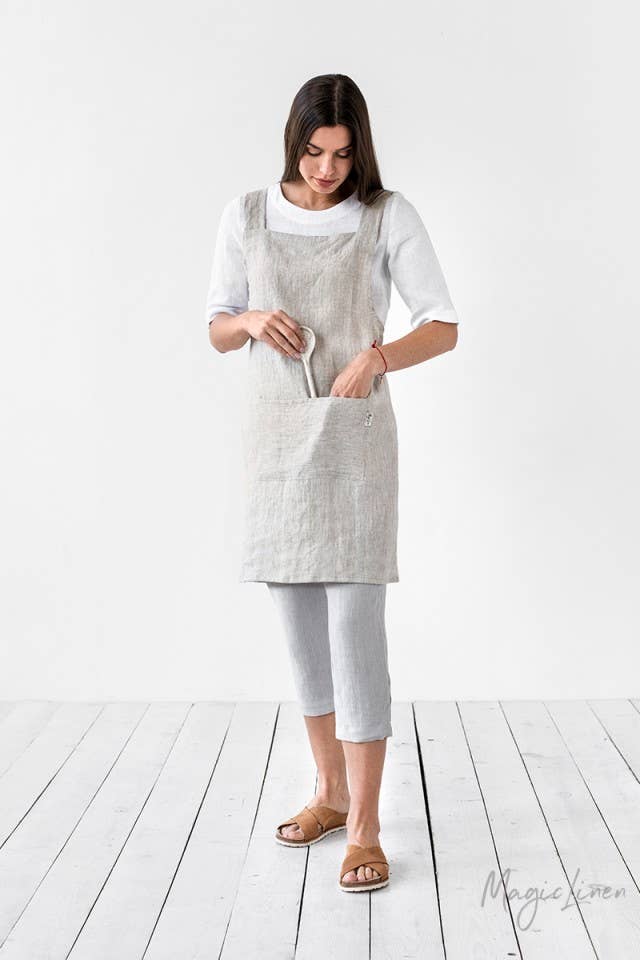 Woman wearing MagicLinen's pinafore cross-back linen apron with twin-sectioned front pocket, ideal for kitchen or garden use, available in various colors.