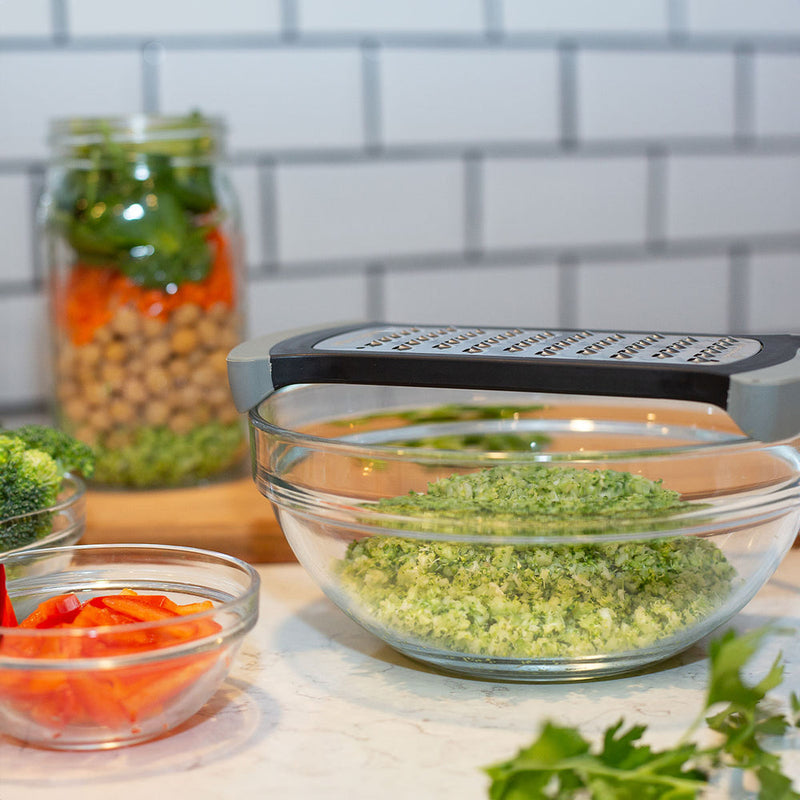 Microplane Bowl Grater on a bowl of grated broccoli, surrounded by sliced peppers, broccoli florets, and a layered mason jar of spinach, carrots, and chickpeas.