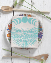 Hearth and Harrow - Organic Cotton Luna Moth Tea Towel with moon phases, displayed on a vintage plate with wooden spoons and wildflowers.