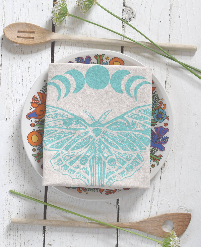 Hearth and Harrow - Organic Cotton Luna Moth Tea Towel with moon phases, displayed on a vintage plate with wooden spoons and wildflowers.