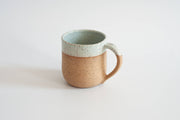 RachaelPots - 12 Oz Original Mug: Sage, hand-thrown ceramic with a thumb rest, featuring a tan base and blue-green glazed rim, perfect for coffee or tea.