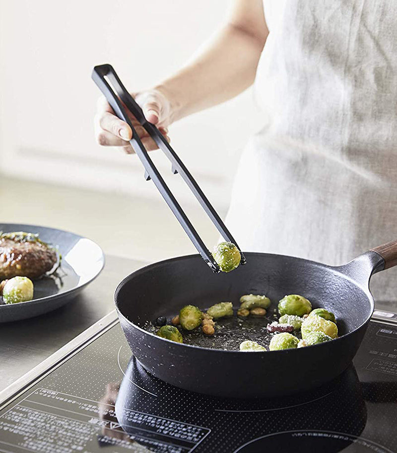 Person uses Yamazaki Home | Tower Floating Tweezer Tongs, Silicone, to lift Brussels sprout from sauté pan on induction cooktop, demonstrating floating design feature.