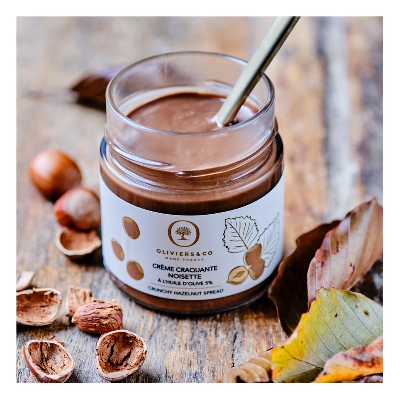 Oliviers&Co | Crunchy Hazelnut Spread in a jar with spoon, surrounded by hazelnuts and shells on a wooden surface.