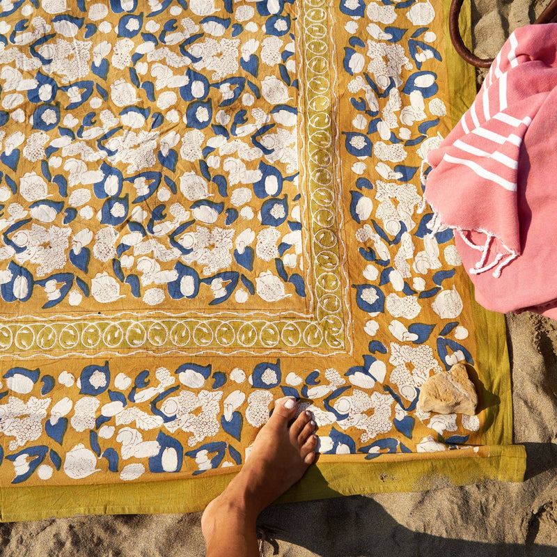 Couleur Nature - Forest Harvest Mustard & Blue tablecloth laid on sand, featuring floral patterns and a small rock, with a foot and striped towel nearby.