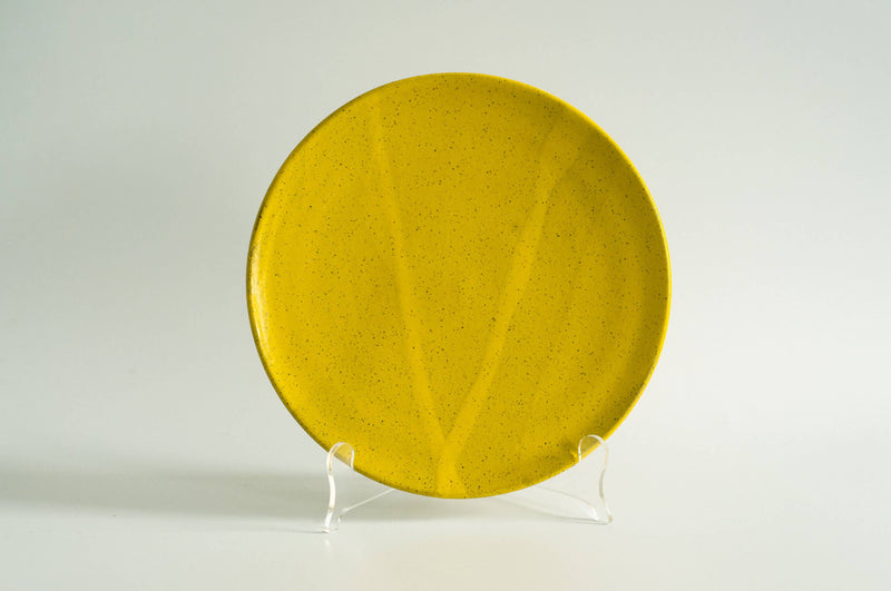 RachaelPots - Dinner Plate: Nude/Light Blue, a round ceramic plate with fine speckles, displayed upright on a clear stand, showcasing its flat middle and upturned lip design.