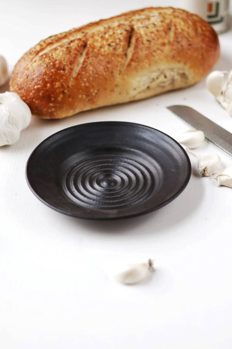 Gravesco Pottery - Garlic Grater Plate in Black with concentric ridges, surrounded by crusty bread and garlic on a minimalist kitchen tabletop.